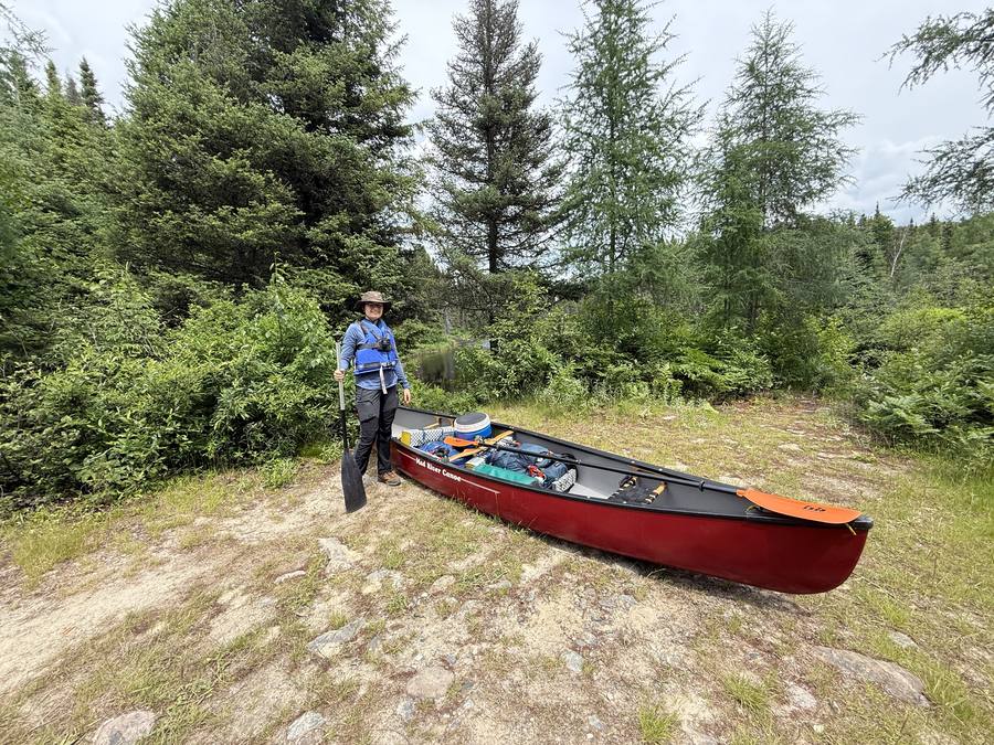 Violette in a sun hat and blue life jacket beside the loaded canoe at the launch, holding a paddle.