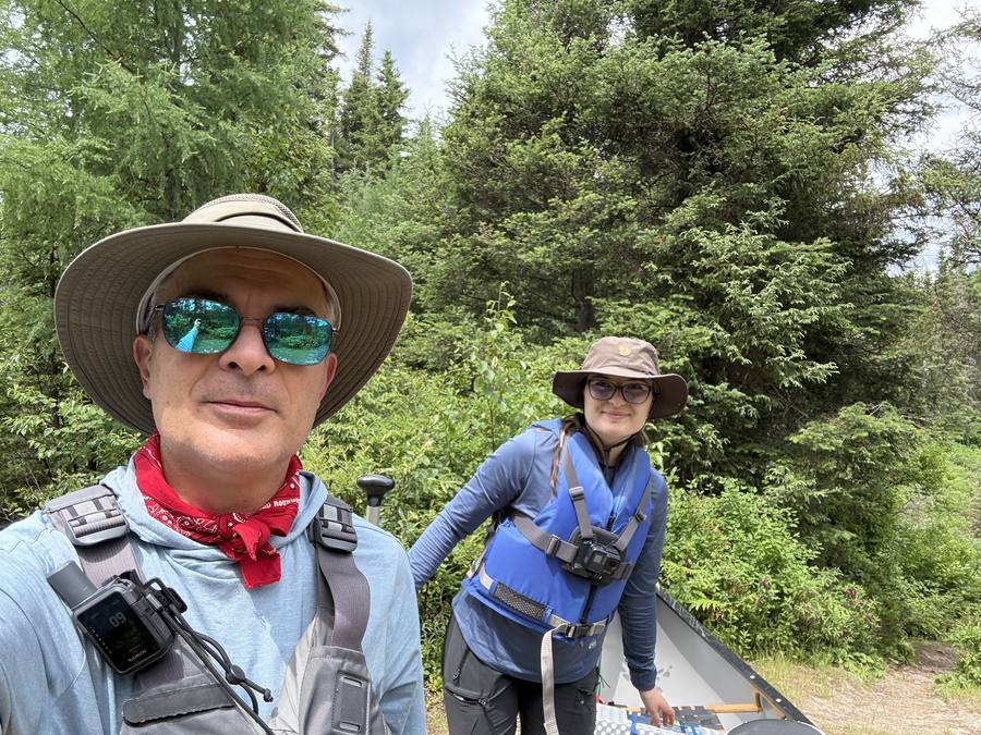 Stan and Violette in hats and life jackets at the launch, forest behind them.