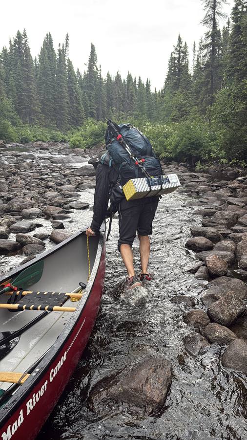 Stan wading down a rocky stream leading the red Mad River canoe by a rope, a large pack on his back.