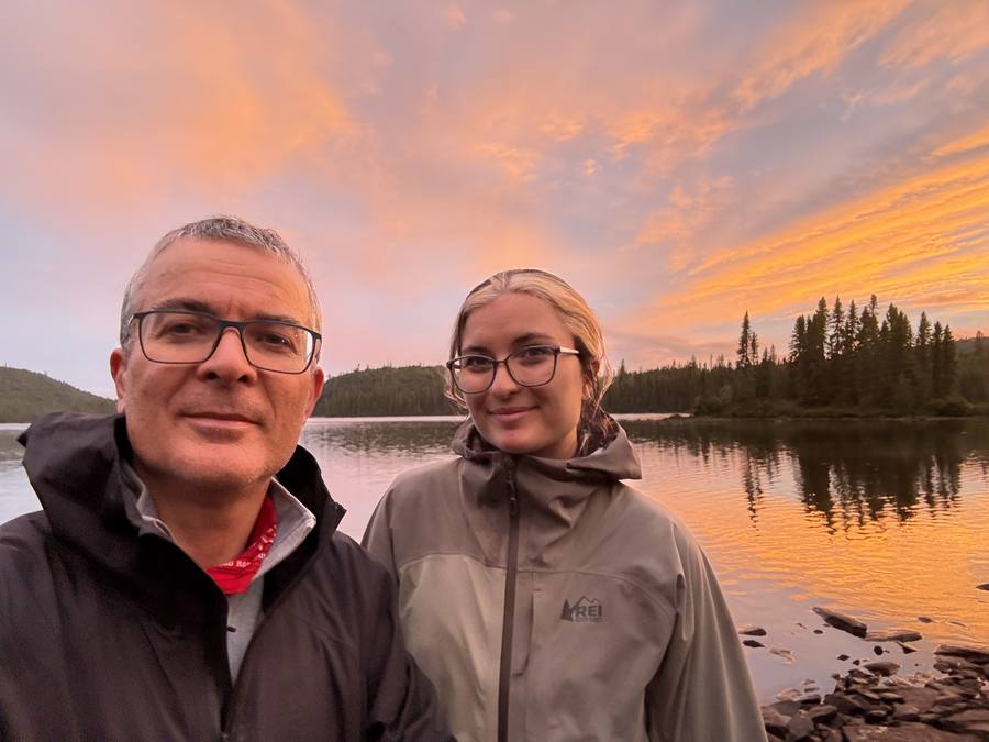 Stan and Violette in rain jackets at camp, a glowing sunset sky reflected on the water behind them.