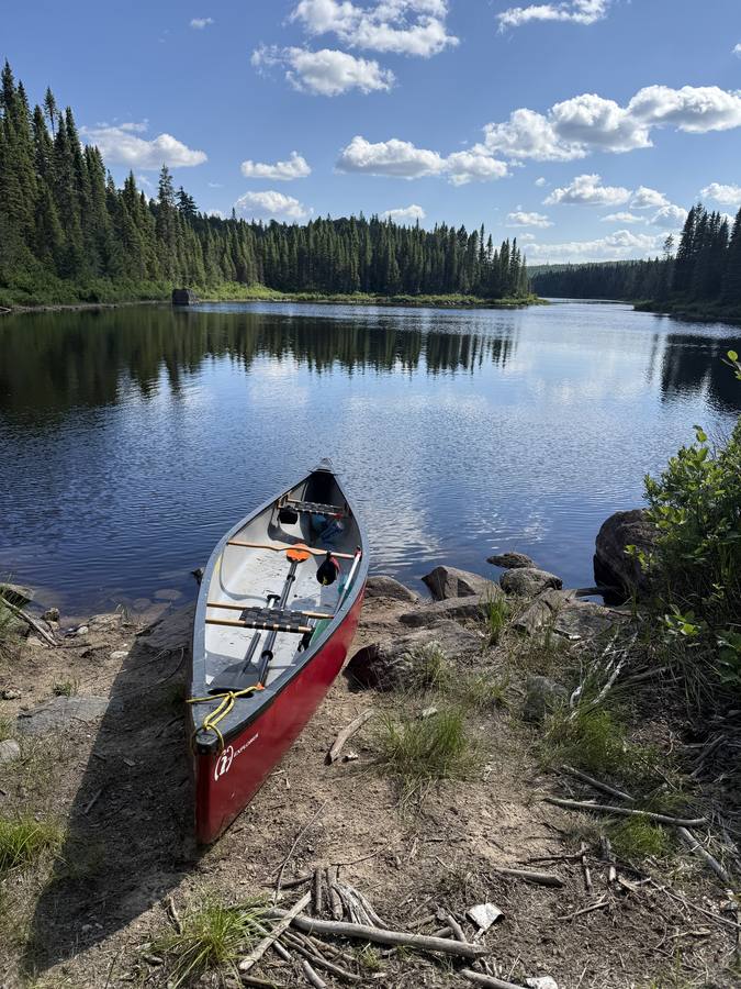 The empty canoe beached on a rocky shore, blue sky and clouds reflected in a calm lake.