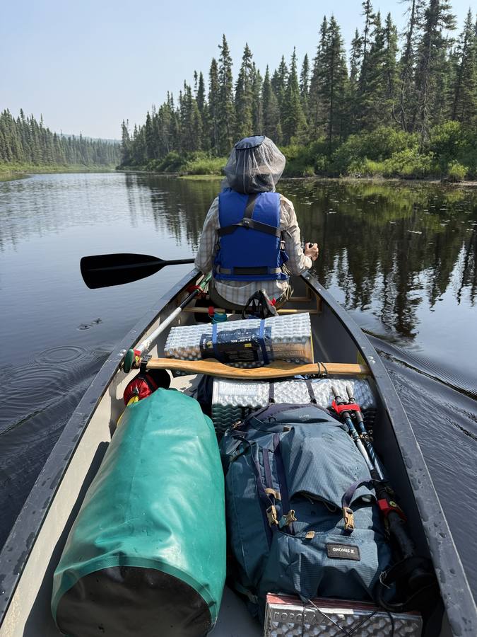 View from the stern of Violette paddling in a bug head net, gear packed in the canoe.