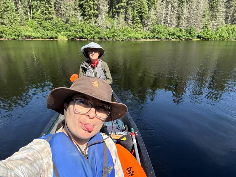 Violette and Stan paddling a calm dark lake, both in hats and life jackets, forest behind them.