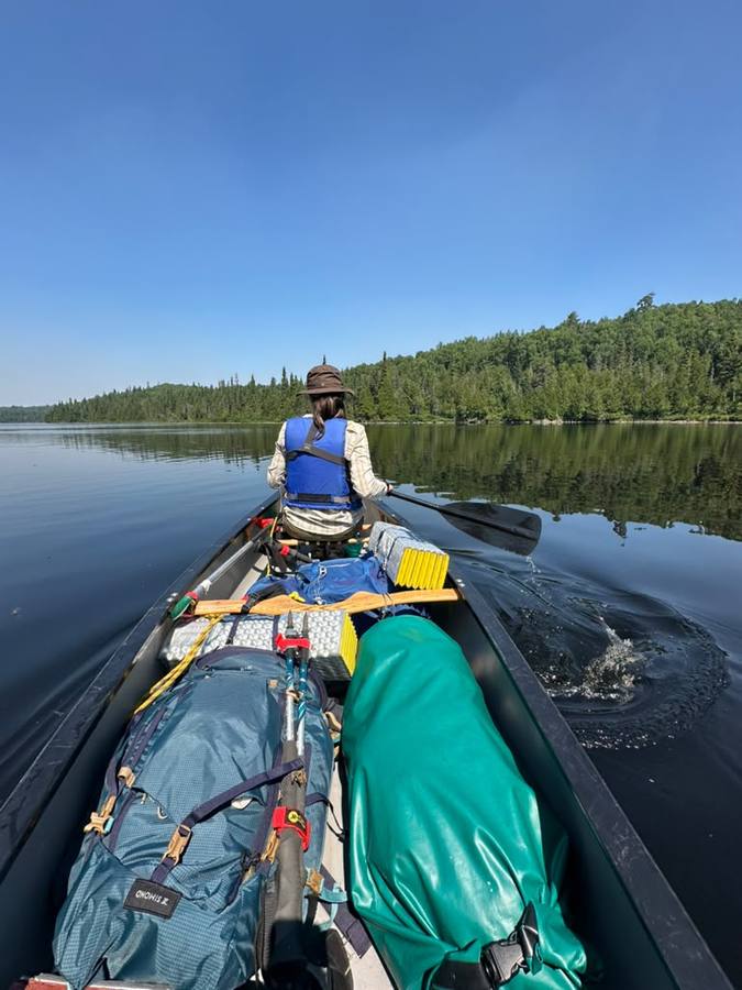 View from the stern of a paddler in the bow crossing a calm blue lake under a clear sky, gear packed in the canoe.