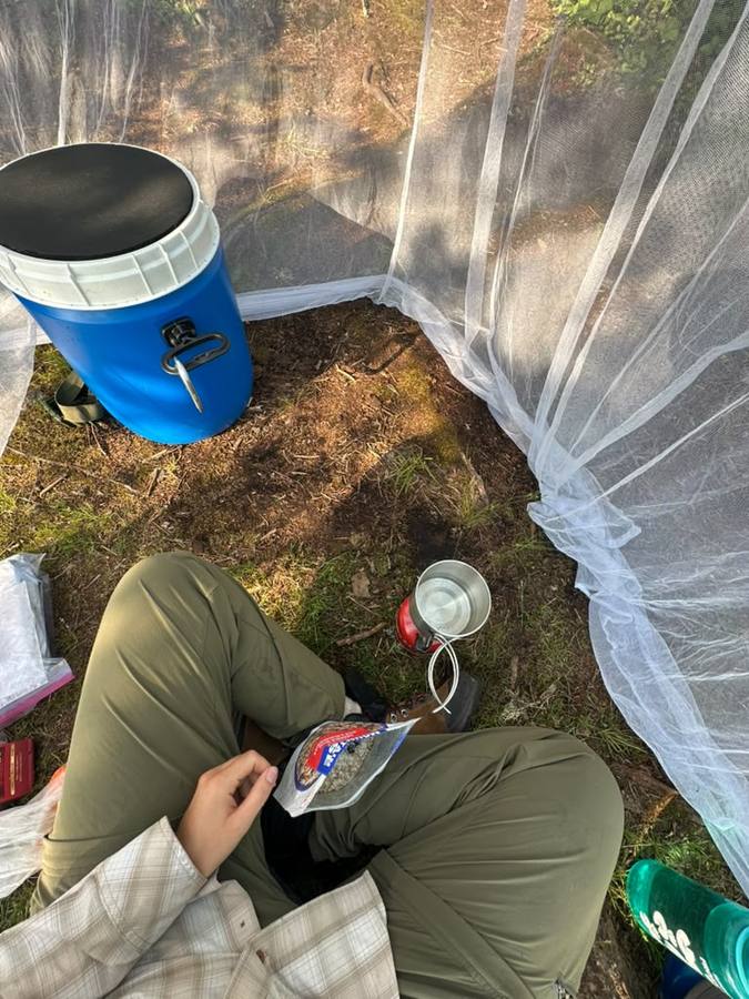 Inside the mosquito shelter: the blue food barrel, a stove, and a freeze-dried meal pouch on a lap.