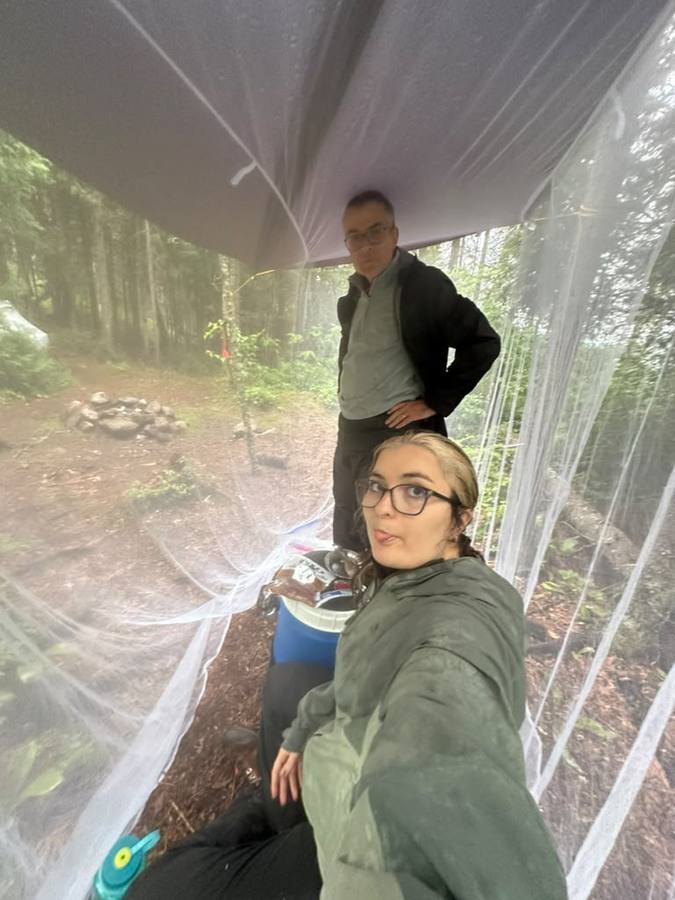Violette and Stan under the mosquito shelter at a wet forest camp, a fire ring outside.
