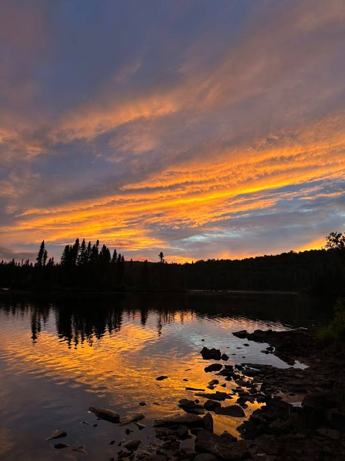 A vivid orange sunset over a calm lake with spruce silhouettes and a rocky shore.