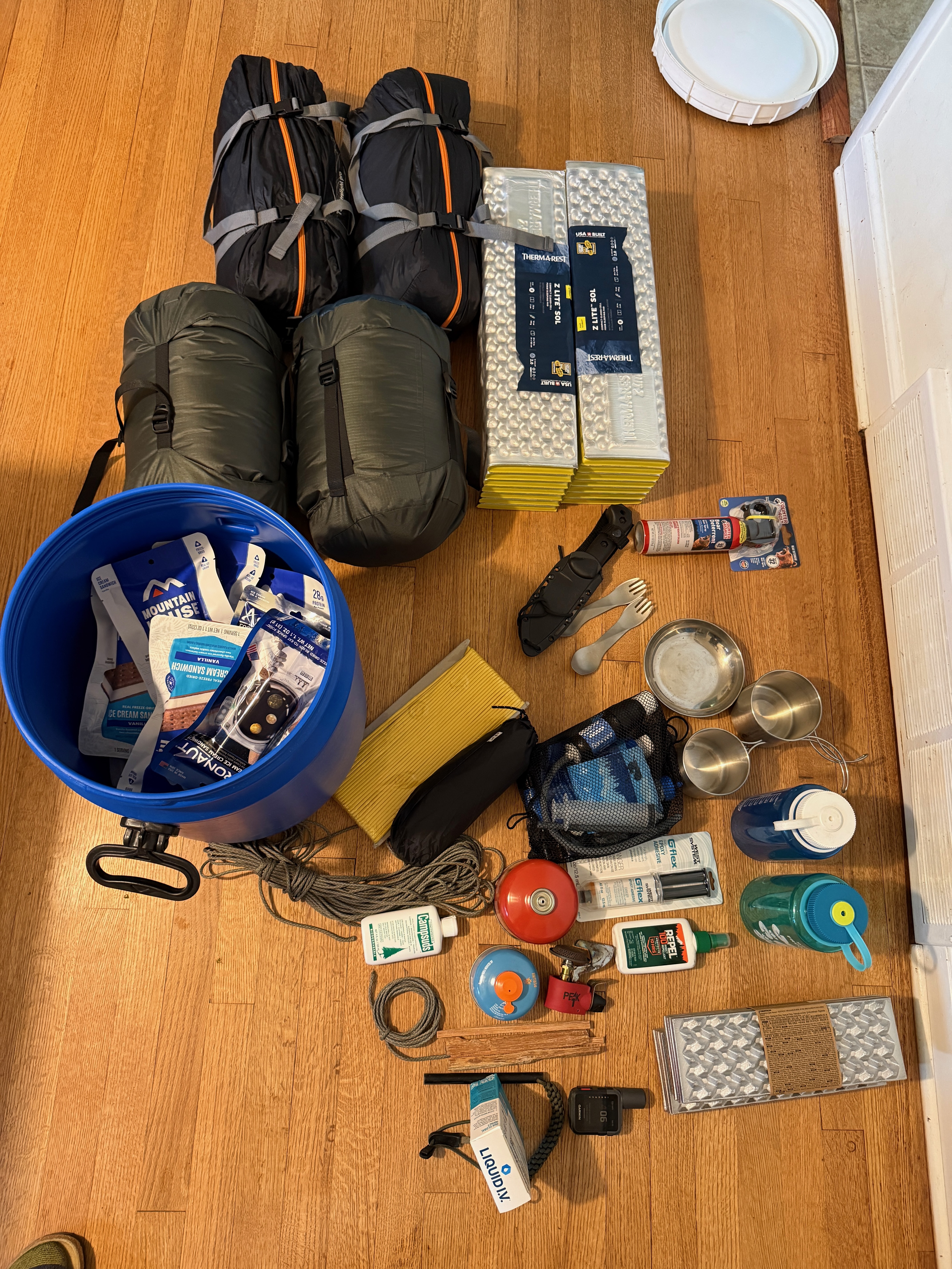 Flat lay of expedition gear: two sleeping bags, two Z Lite SOL pads, blue food barrel, rope, knife, spork, stainless cups and bowls, Nalgenes, fuel canisters, MSR stove, insect repellent, and Liquid IV.